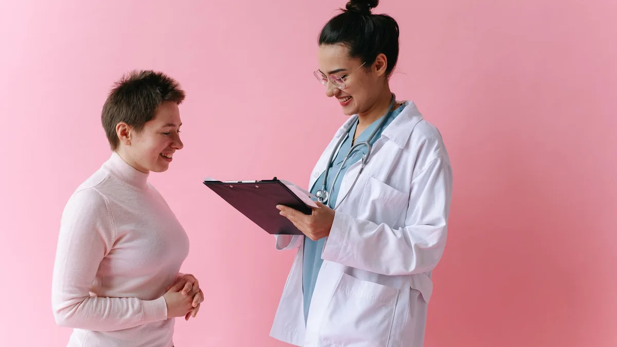 A doctor in a white coat consults a smiling patient against a pink backdrop.
