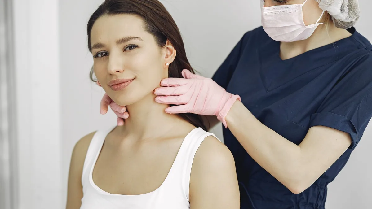A medical practitioner in scrub suit performs a neck examination on a smiling woman in a clinic.