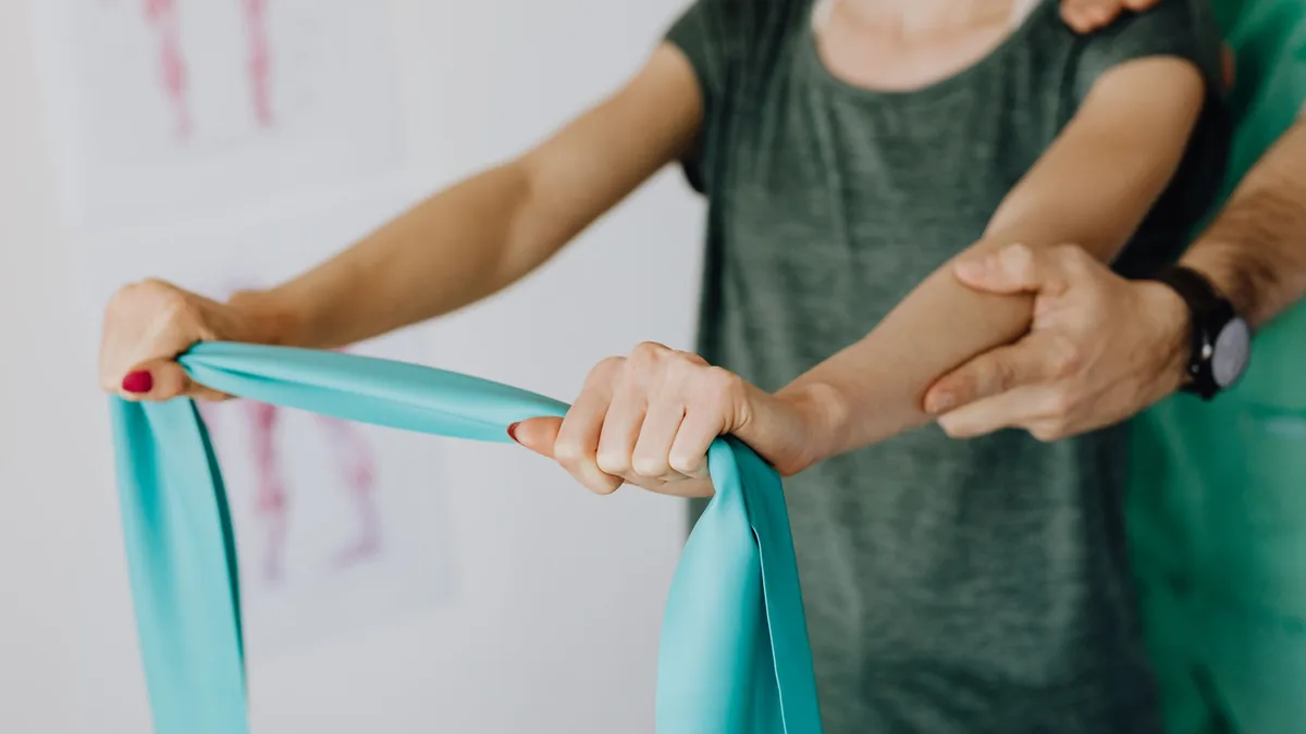 Crop unrecognizable orthopedist in uniform and wristwatch supporting shoulder and reached arm of faceless female patient with resistance band in.