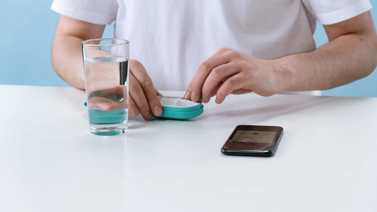 A person arranging pills with a weekly organizer, next to a glass of water and a smartphone.