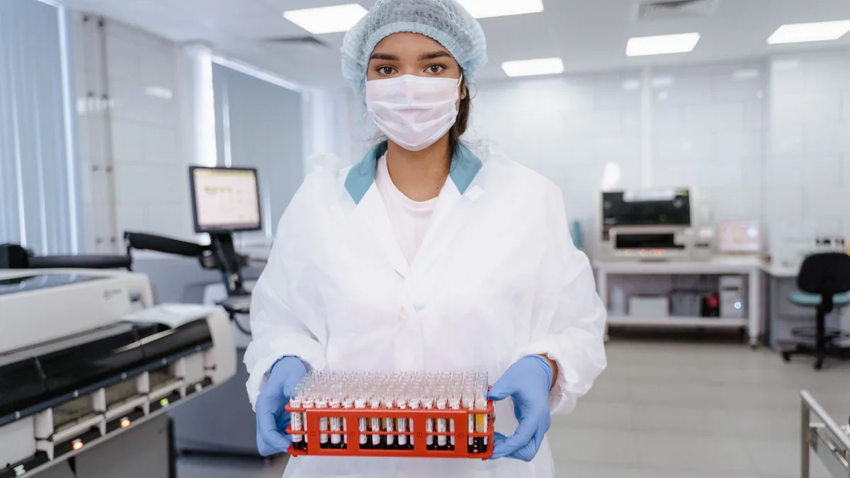 Lab technician in protective gear holding a tray of test tubes in a modern laboratory setting.