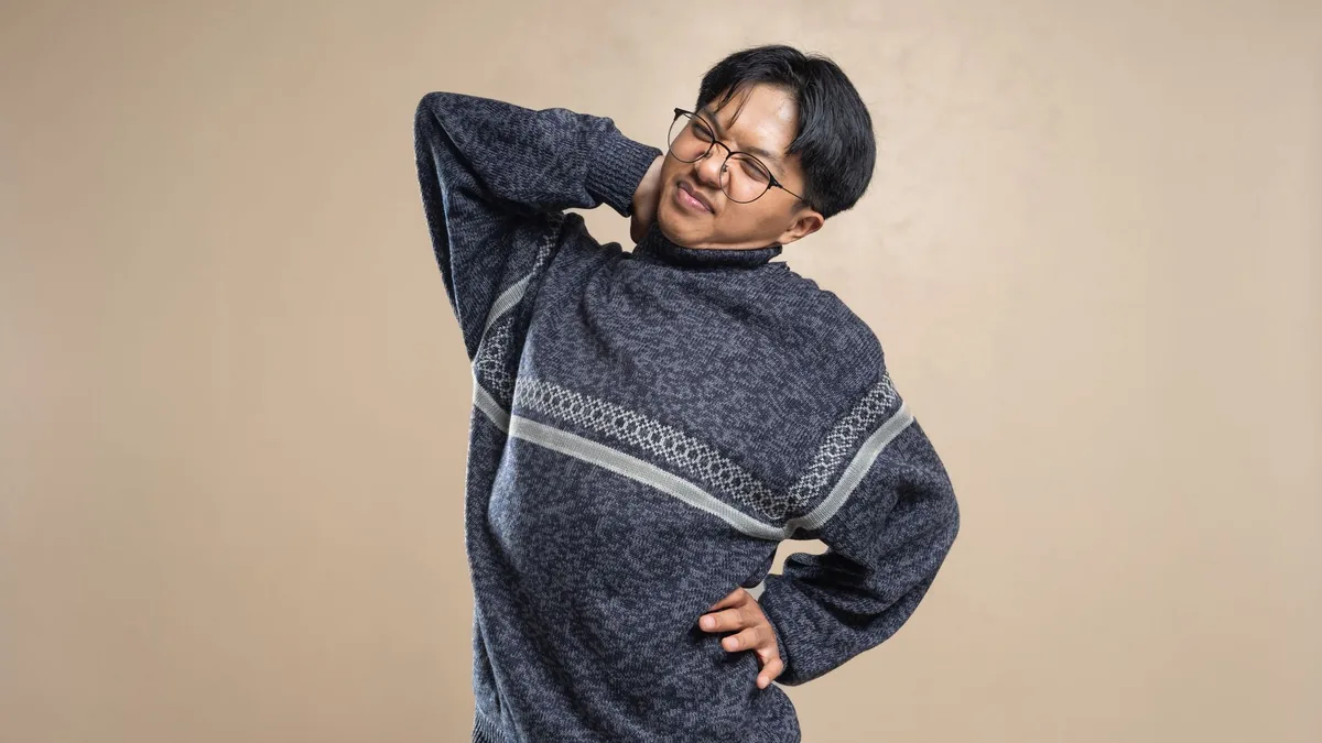 Asian man wearing eyeglasses and sweater stretching indoors, feeling body pain.