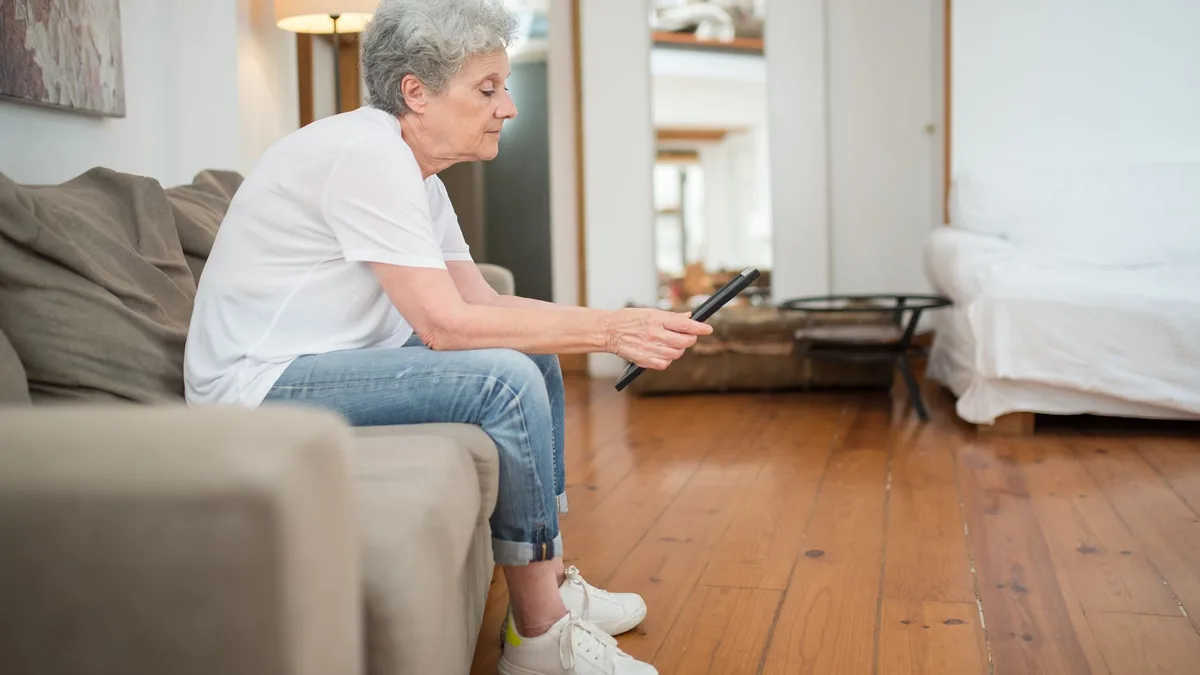 Senior woman in casual attire sitting on a couch holding a tablet in a cozy living room.