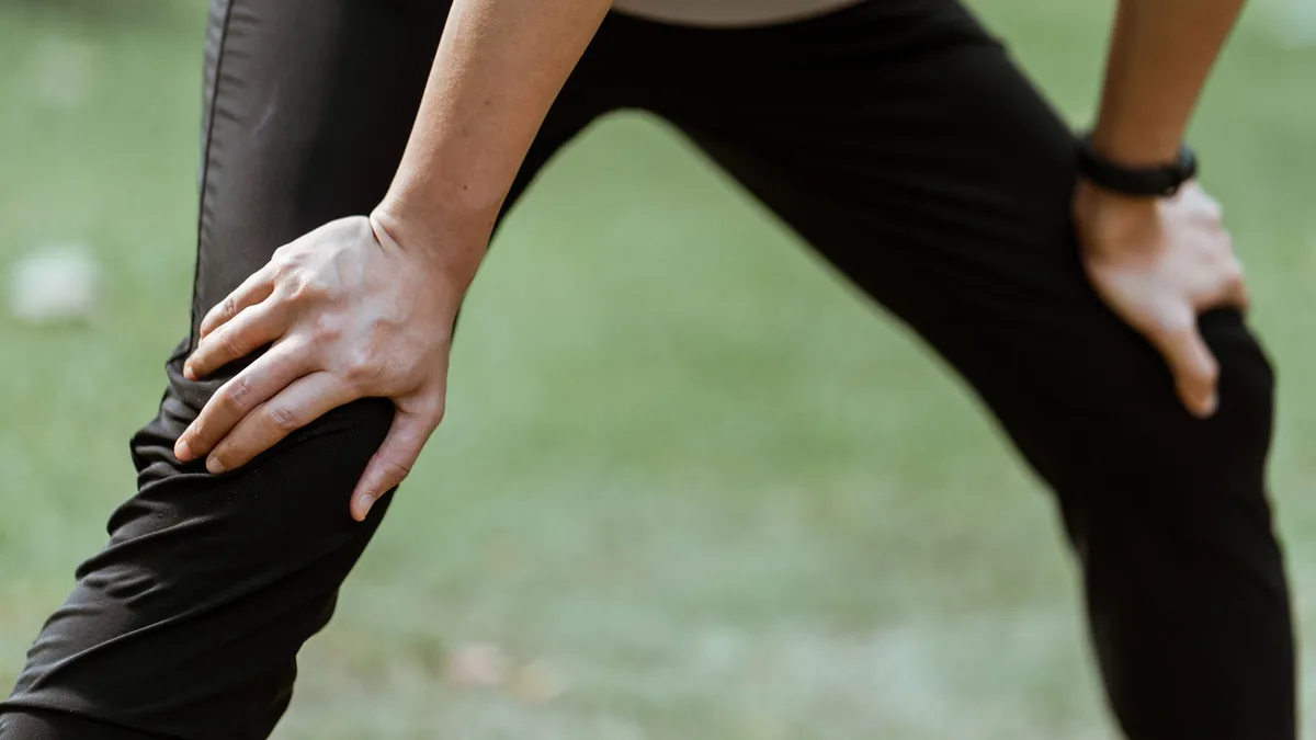 Crop unrecognizable sportsperson standing on recovery posture with hands on knees after active workout in green park.