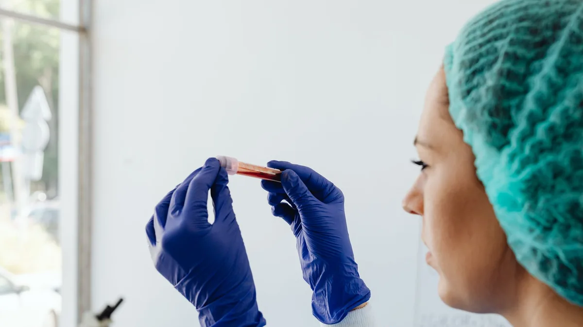 A focused lab technician holds a blood sample in a test tube, wearing gloves and a hair net.