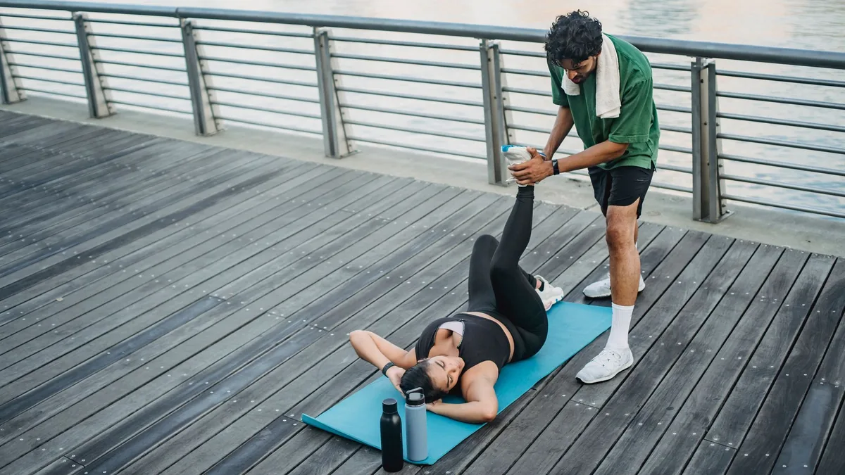 Man assisting woman in stretching exercises on boardwalk next to river.