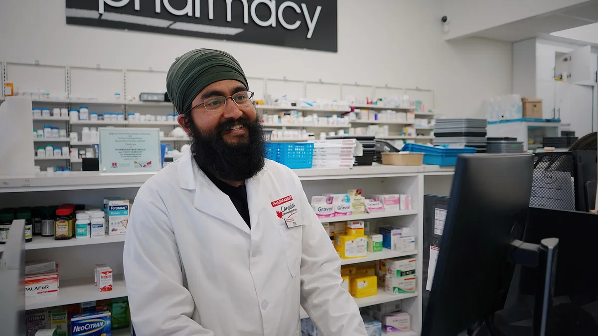 Male pharmacist with a turban standing and smiling in a pharmacy, working at the counter.