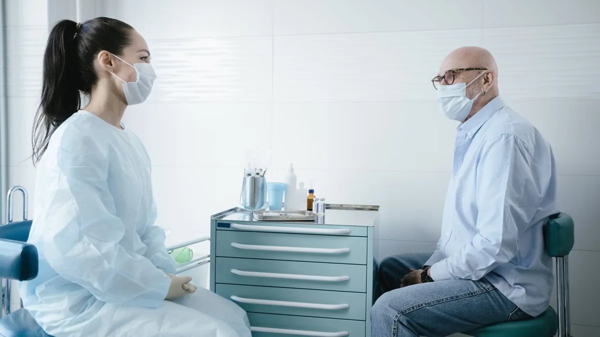 Medical professional consults a senior patient, both wearing masks in a clinic setting.
