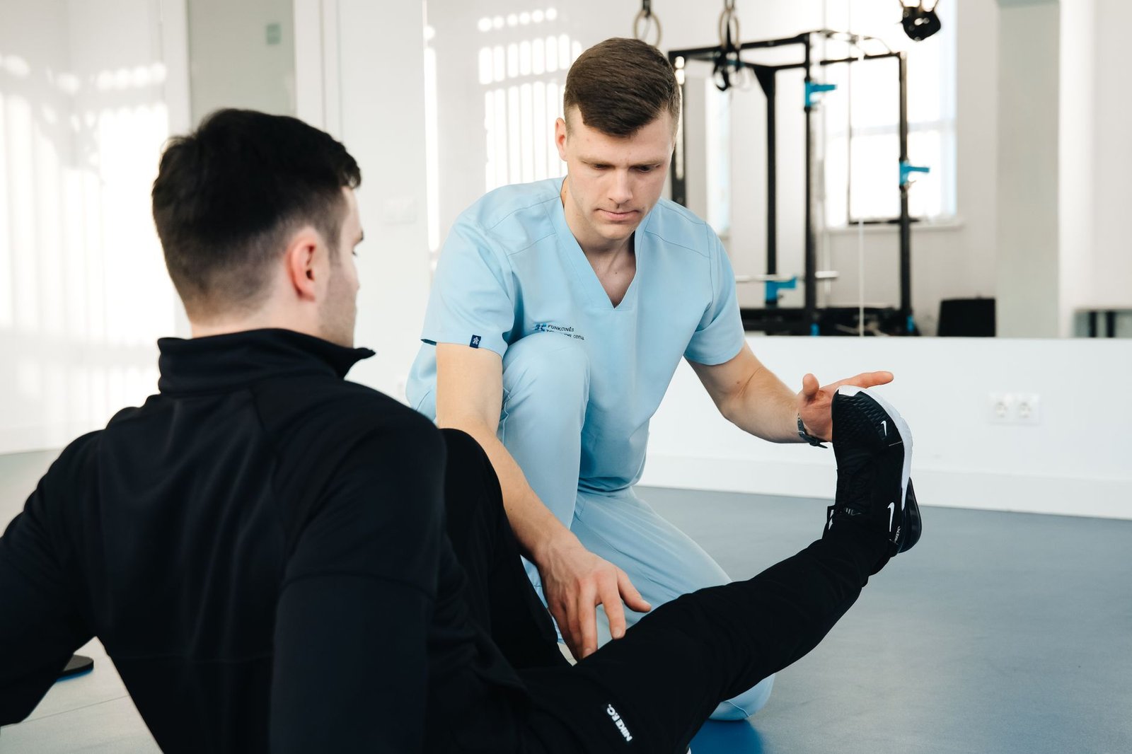 A therapist in scrubs guiding a seated man through a straight-leg exercise in a rehabilitation gym
