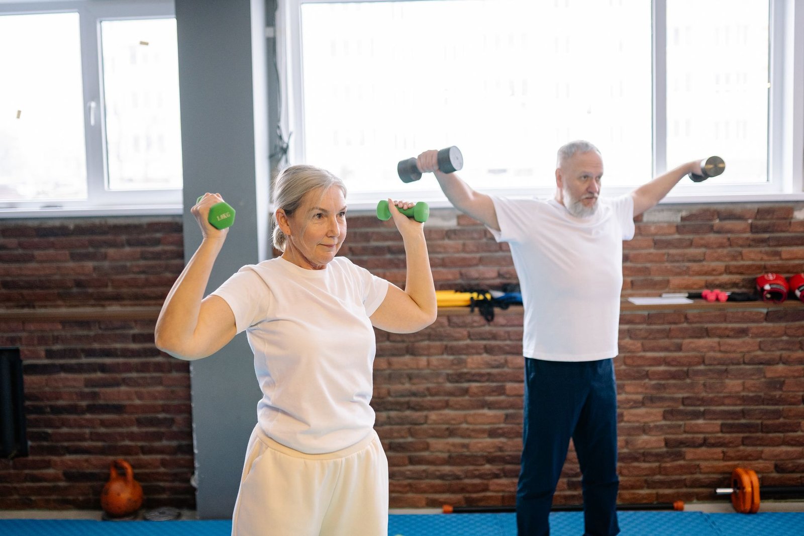An older woman and an older man performing dumbbell shoulder exercises side by side in a studio