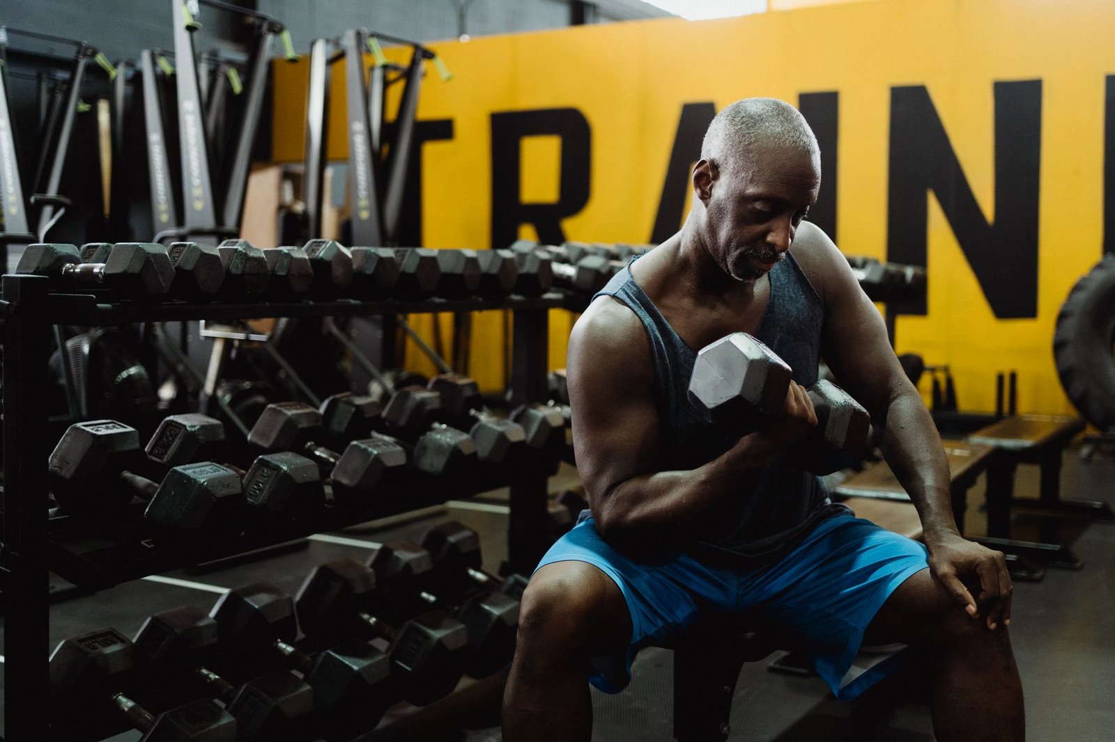 A grey-haired man seated beside a dumbbell rack performing a controlled biceps curl in a gym
