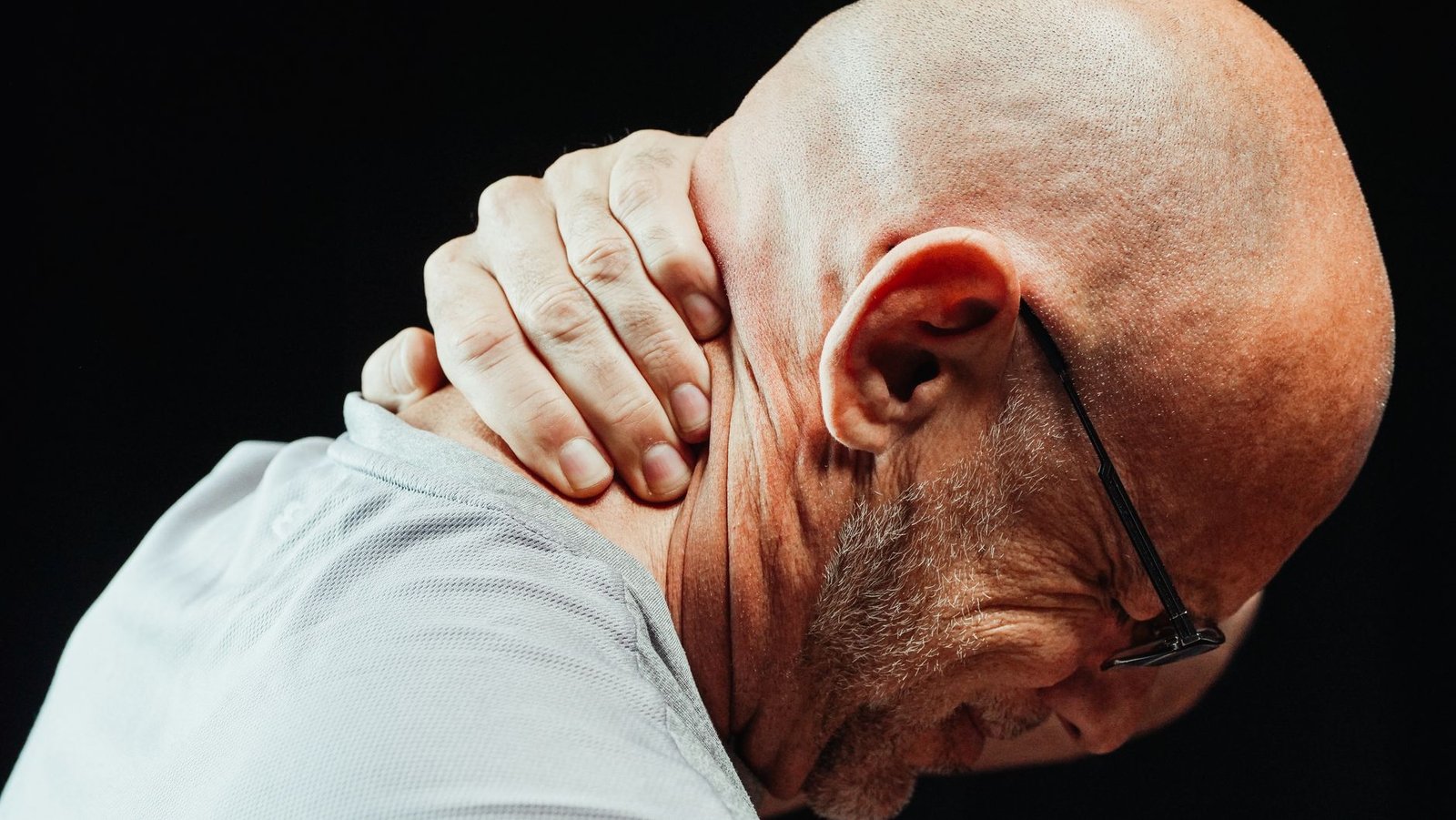 An older man in glasses gripping the back of his neck with his head bowed, against a dark background