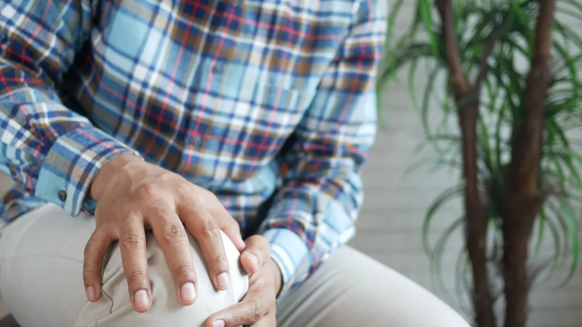 Seated man in a plaid shirt holding his painful knee with both hands