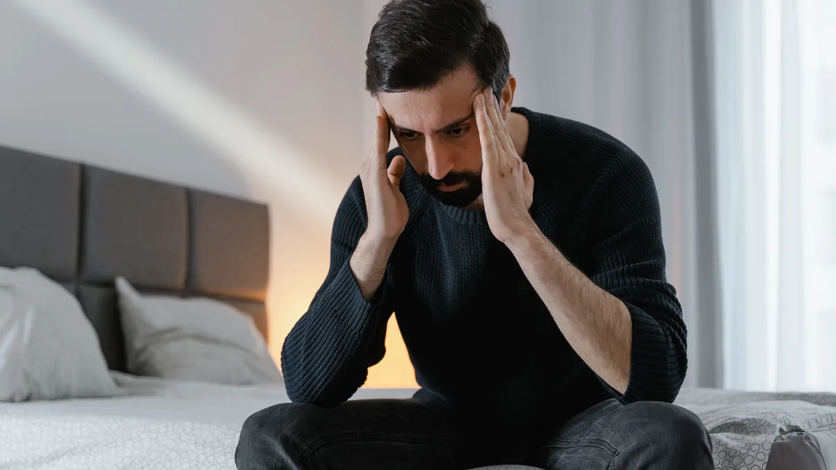 Man sitting on the edge of his bed with his head in his hands, worn down by pain that has not resolved