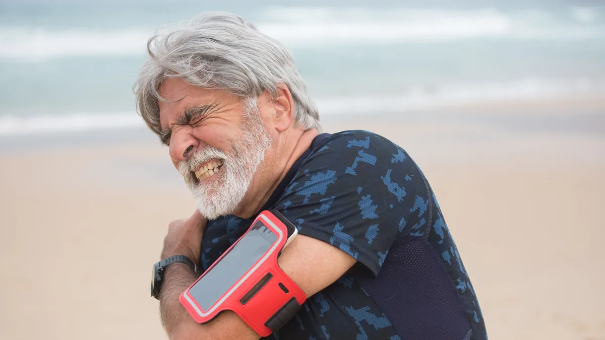 Older man grimacing and holding his painful shoulder on a walk by the beach