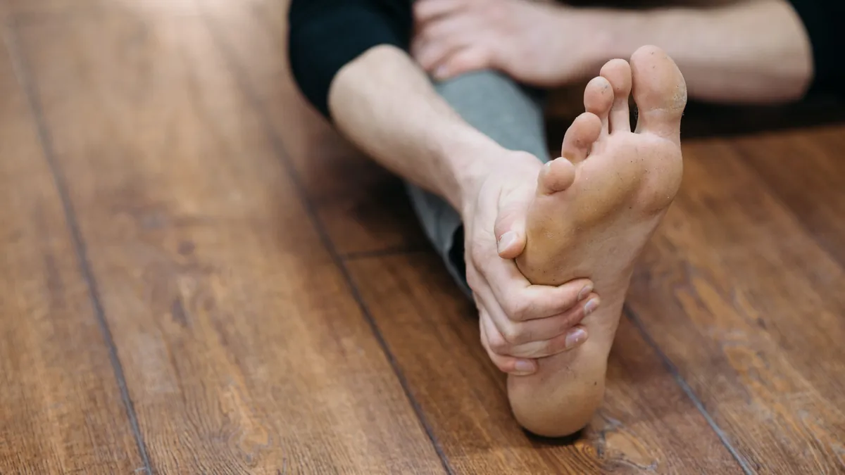Person seated on a wooden floor stretching the sole of a bare foot with one hand