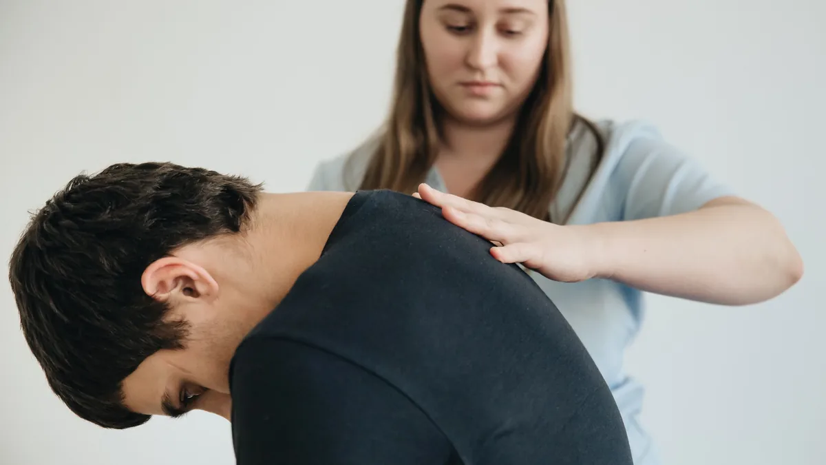 Physical therapist placing a hand on the upper back of a man bent forward with back pain