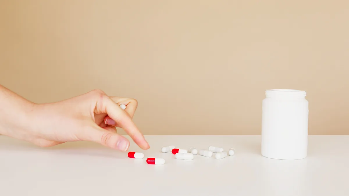 Hand picking up one pill from a few scattered on a table beside a medicine bottle